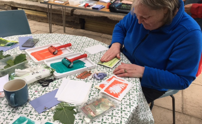 A woman in a blue jumper folders a piece of paper in half. She is sitting at a table covered in printing materials - blocks and trays of green and red paint