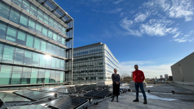 Two people stand next to solar panels on the rooftop of the Unicorn Theatre in London. It is a sunny day and the light is catching the nearby glass building