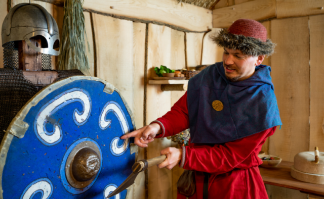 An actor dressed as a Viking at the JORVIK Centre in York points at a blue circular shield in front of a suit of armour