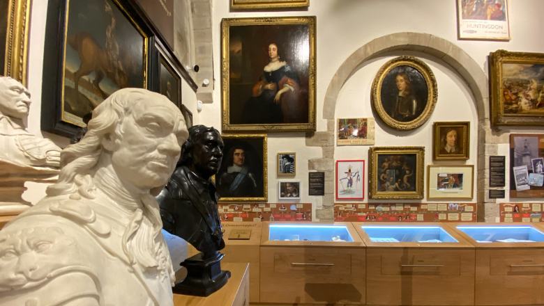 The image shows a museum display. The foreground shows the marble bust of a man with long hair, whilst the background shows a wall with oil paintings and a display case with objects inside.