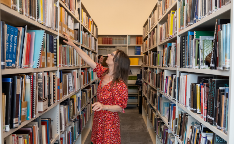 A woman in a red dress reaches up to a bookshelf in the Wolfson Library at the Royal Academy of Dance