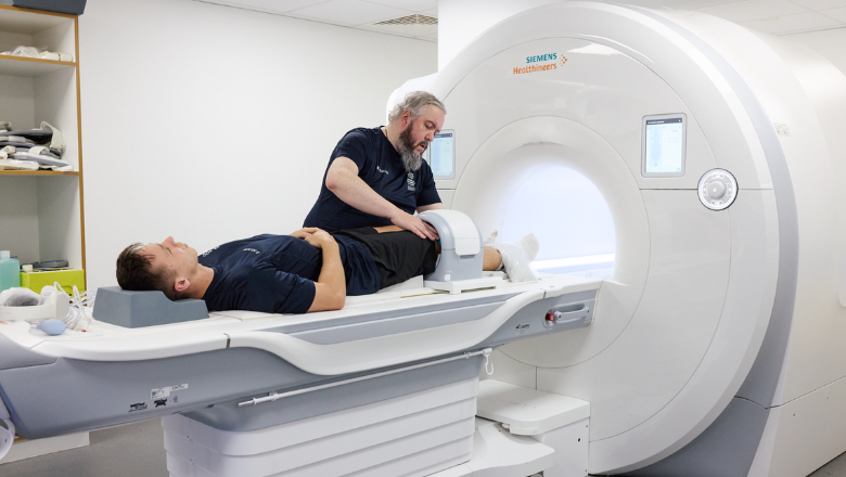 A patient lies on a bed while a man prepares an MRI scanner for the test.