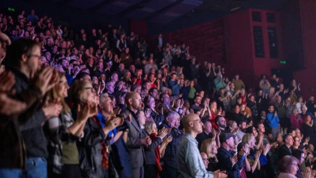 A full audience give a standing ovation and clap a performance on stage.