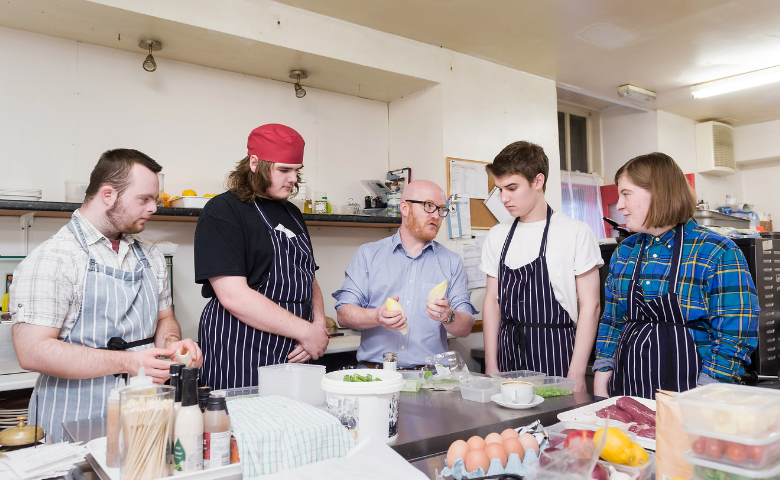 Giraffe training kitchen. Five people stand in a kitchen with food in front of them. One, a teacher holds an ingredient while the other four listen.