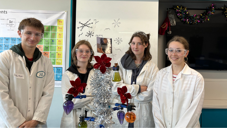 Four students pose with a science experiment in a classroom. The experiment is Christmas-themed, with tinsel.