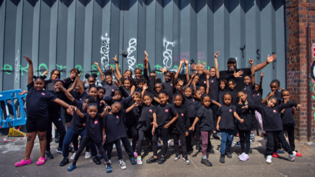 A large group of children celebrate in front of large grey metal doors.