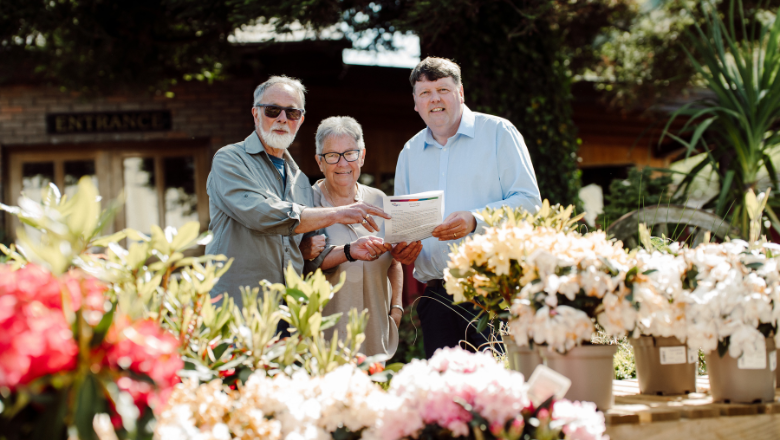 Three people stand outside in the sun holding a piece of paper. There is a table filled with pots of flowers in front of them.