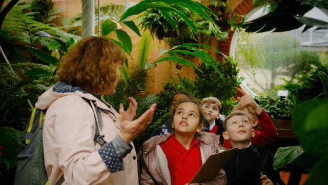 A small group of children stand in a leafy green inside space, looking up and around. An adult gestures with their hands as if speaking to the group.