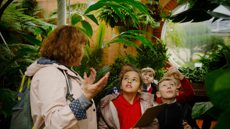 A small group of children stand in a leafy green inside space, looking up and around. An adult gestures with their hands as if speaking to the group.